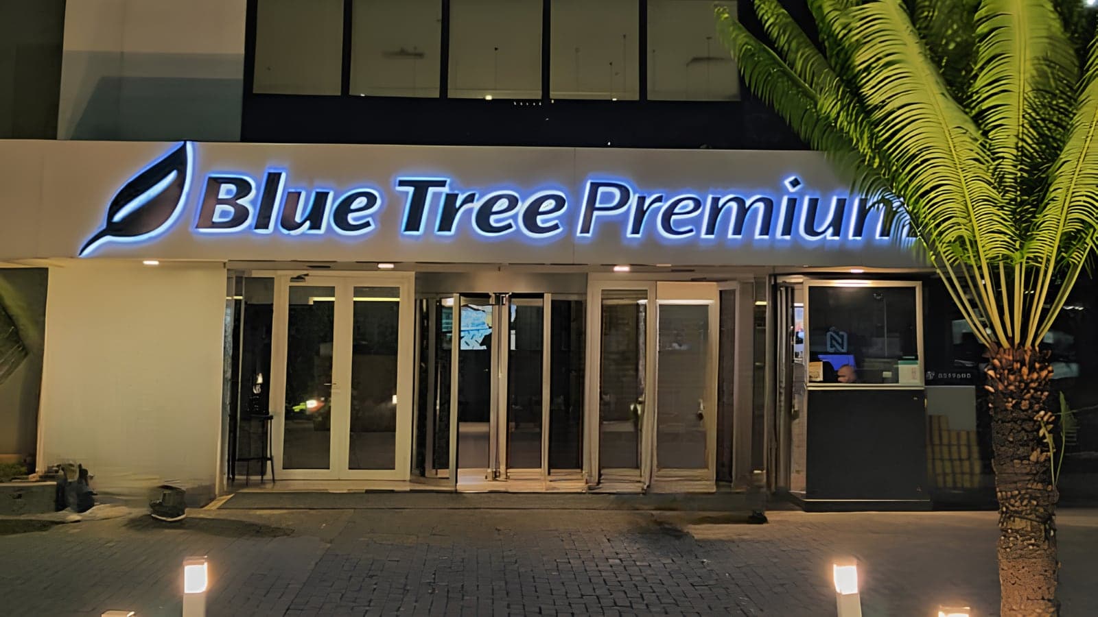 Fachada do Blue Tree Premium Alphaville à noite, com o letreiro iluminado em azul acima da entrada de vidro e uma palmeira à direita.